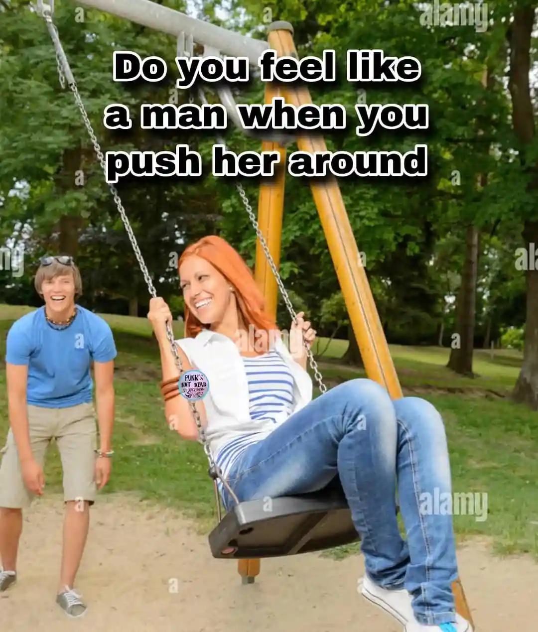 A man in a blue shirt and khaki shorts stands smiling beside a swing set, pushing a woman with red hair who is seated on the…