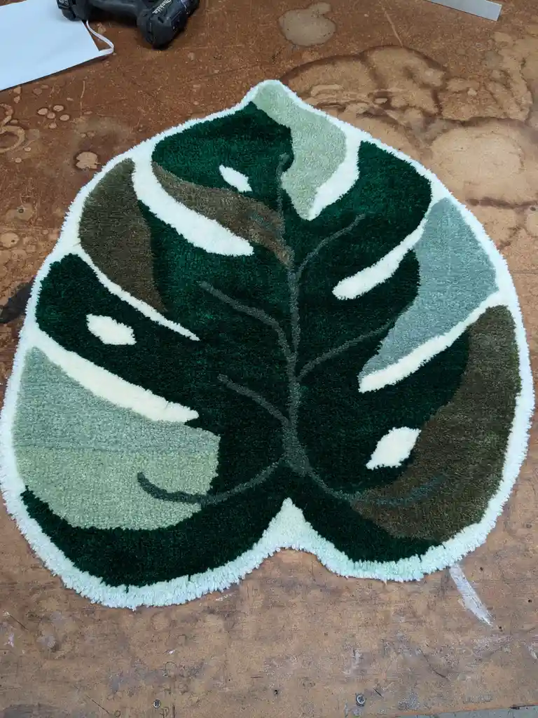 A shaggy, leaf-shaped rug with green, white, and gray sections resembling a monstera leaf lies on a worn wooden workbench…