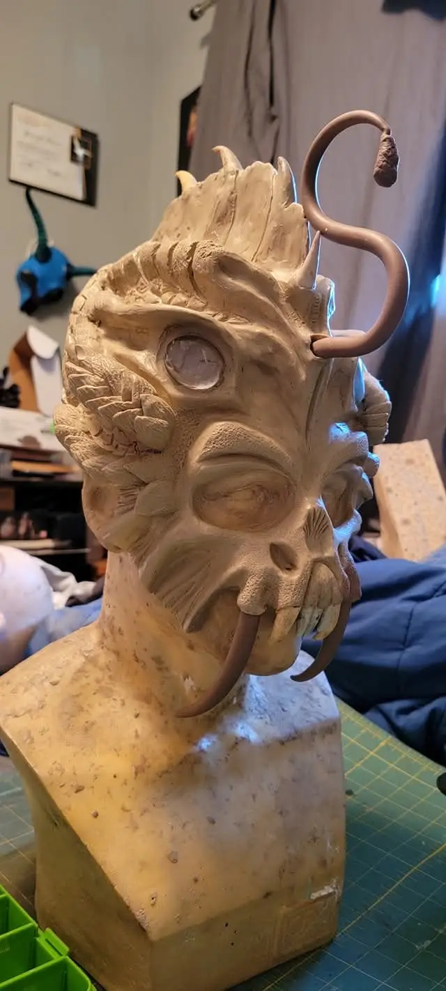 A detailed, unfinished clay sculpture of a demonic head with horns, tusks, and a curled tail-like appendage sits on a…