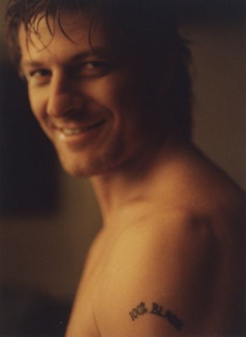 A shirtless man with wet hair smiles at the camera, showing a tattoo on his left shoulder that reads '100% BLAME'. The image…