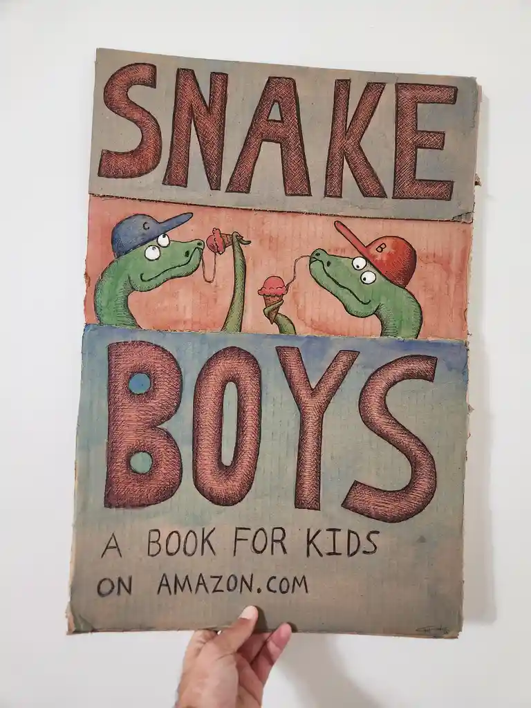 A hand holds a handmade cardboard book cover featuring two cartoon green snakes wearing baseball caps, sharing an ice cream…