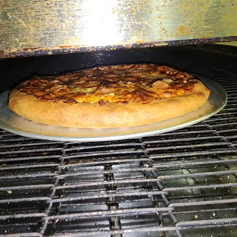A close-up photo shows a round pizza with a golden-brown crust and slightly charred cheese and toppings, resting on a metal…