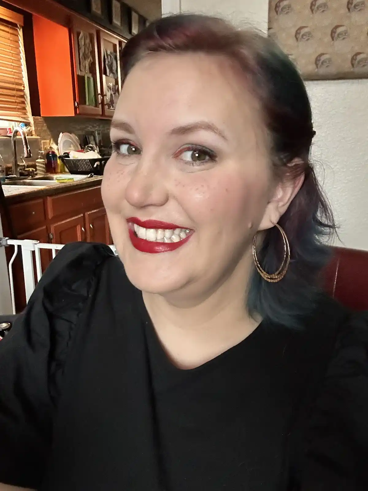 A woman with colorful hair and bright red lipstick smiles while looking off-camera, with a kitchen and a Santa-themed…