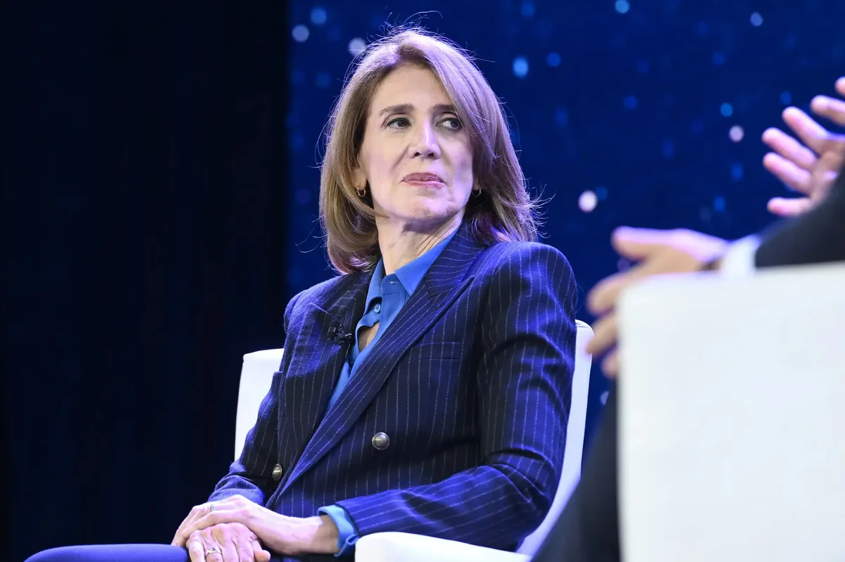 A woman in a pinstripe blazer sits in a white chair, looking sideways with a skeptical expression, while another person's…