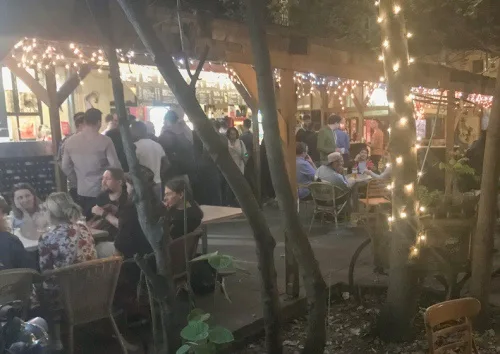 A nighttime outdoor patio scene with people seated at tables under string lights and wooden beams, some standing and…