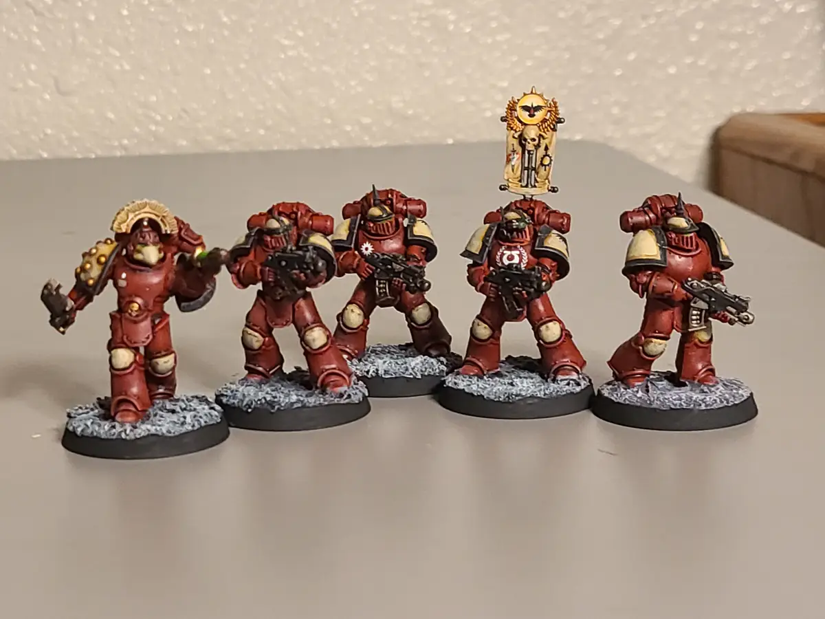 Five painted miniature figures in red and gold armor stand in a row on a flat surface. The figure on the far right has a…