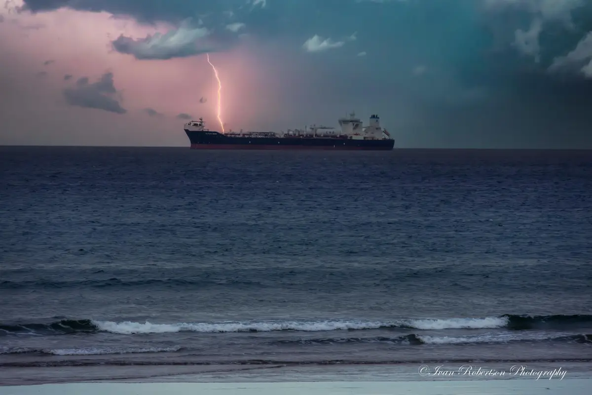 A large LNG tanker ship is seen on the open ocean during a storm, with a bright lightning bolt striking directly above it…