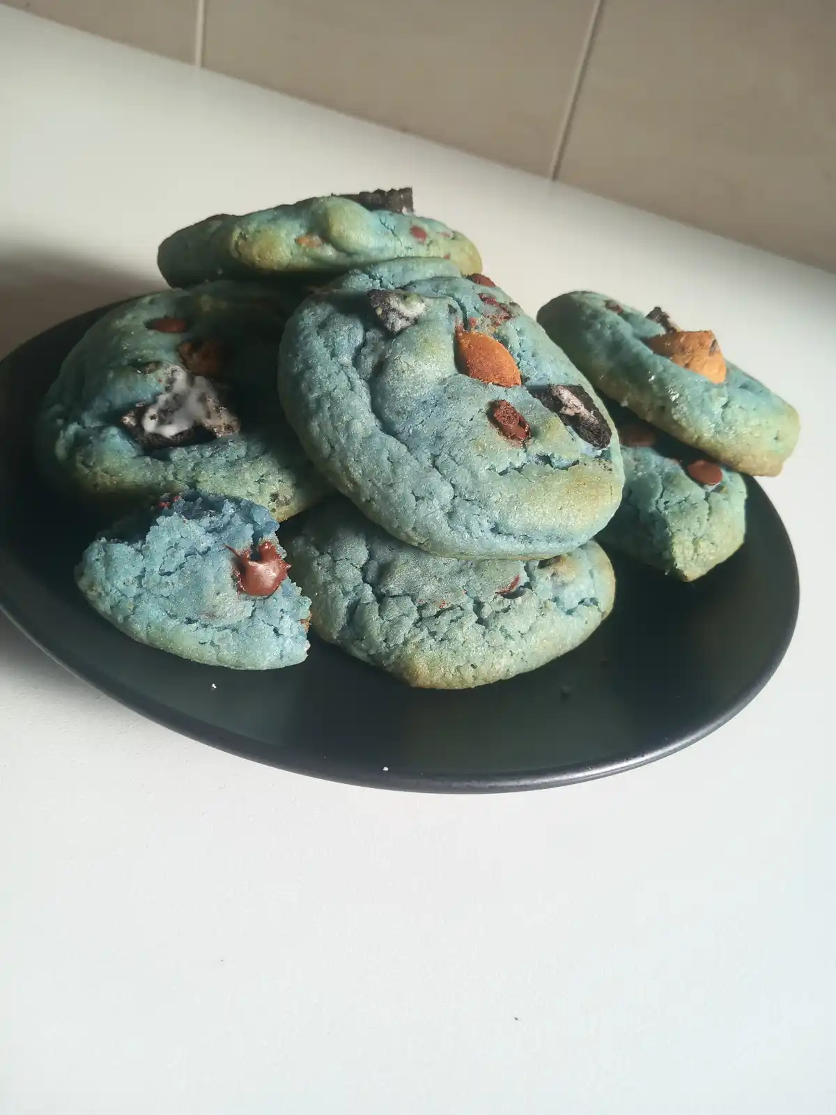 A pile of blue-colored cookies with chocolate chips and Oreo pieces sits on a black plate atop a white countertop. The…