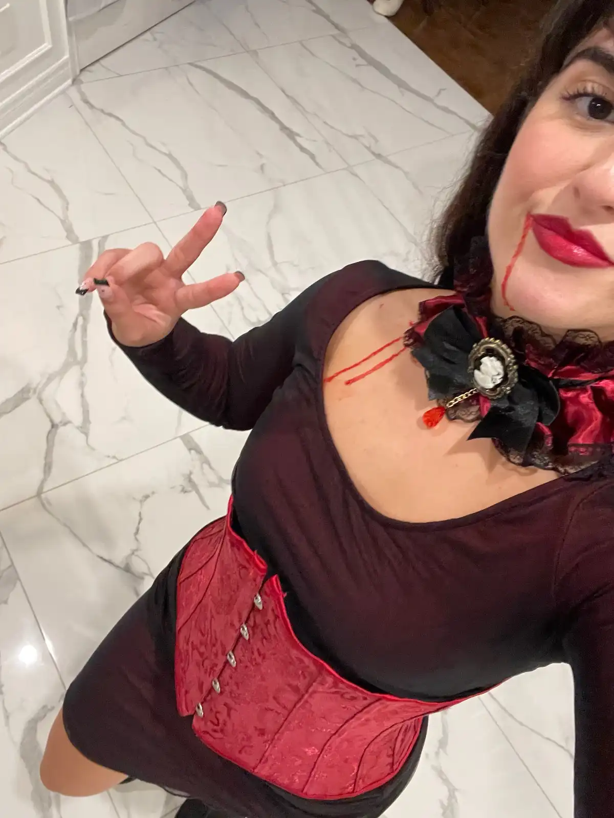 A person in a gothic vampire costume with red lipstick, fake blood dripping from their chin and across their chest, wearing…