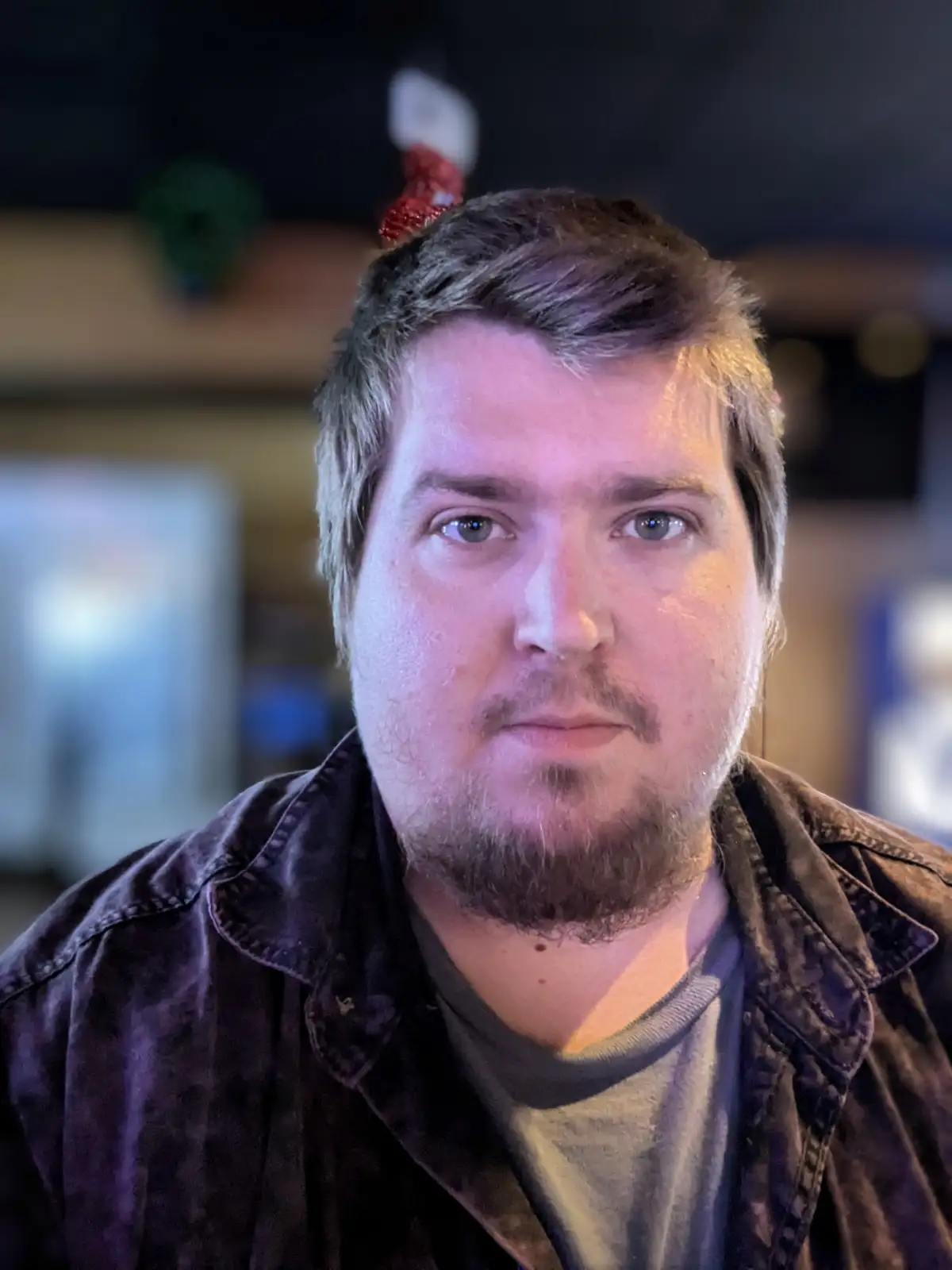 A man with a neutral, slightly unimpressed expression stares directly at the camera, wearing a dark jacket over a t-shirt. A…