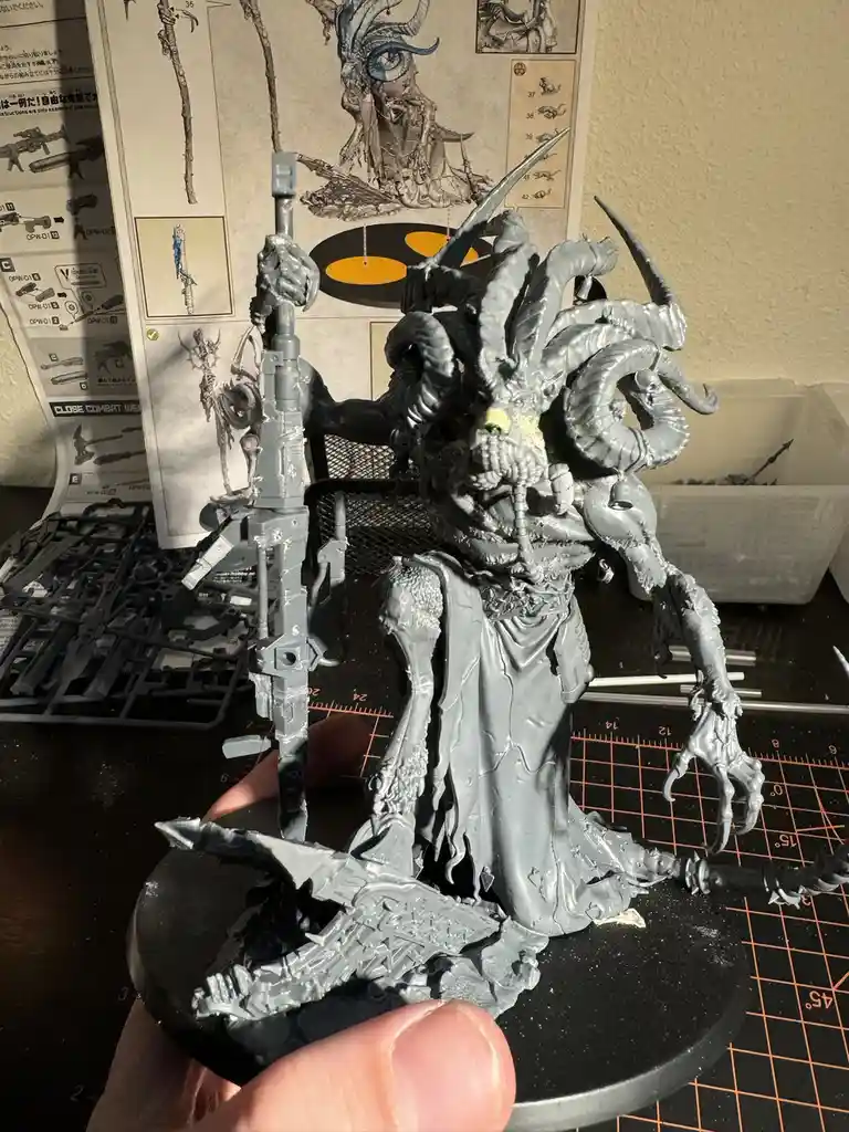 A person holds up a freshly painted, grey miniature of a monstrous, horned demon with multiple arms and weapons, standing on…