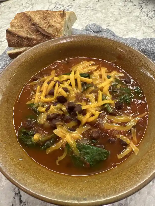 A close-up photo shows a brown ceramic bowl filled with a reddish-brown soup containing black beans, green leafy vegetables…