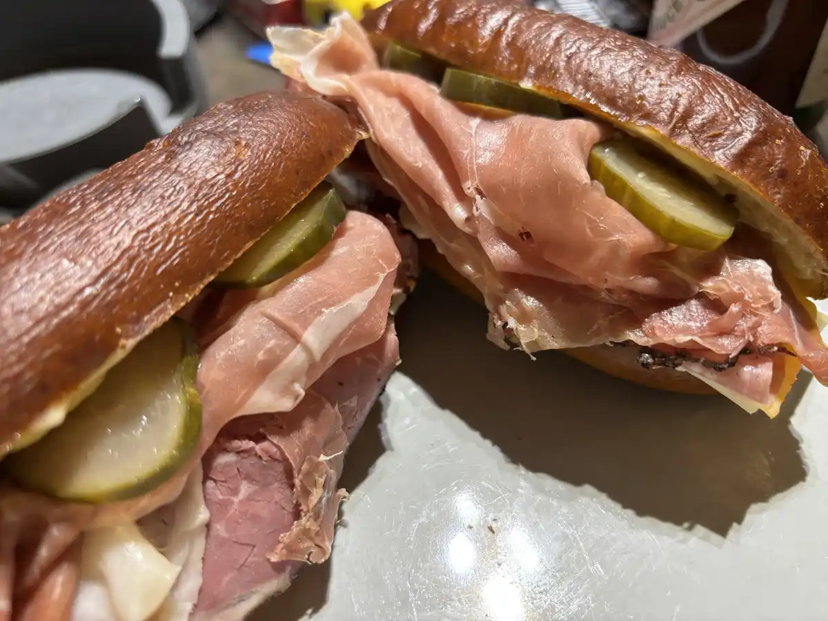 A close-up photo shows a large, overstuffed sandwich cut in half, revealing layers of pink deli meat, pickles, and cheese…
