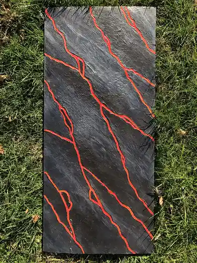 A rectangular black stone slab with bright red, wavy lines painted on it, resembling lava flows, lies on a bed of green…