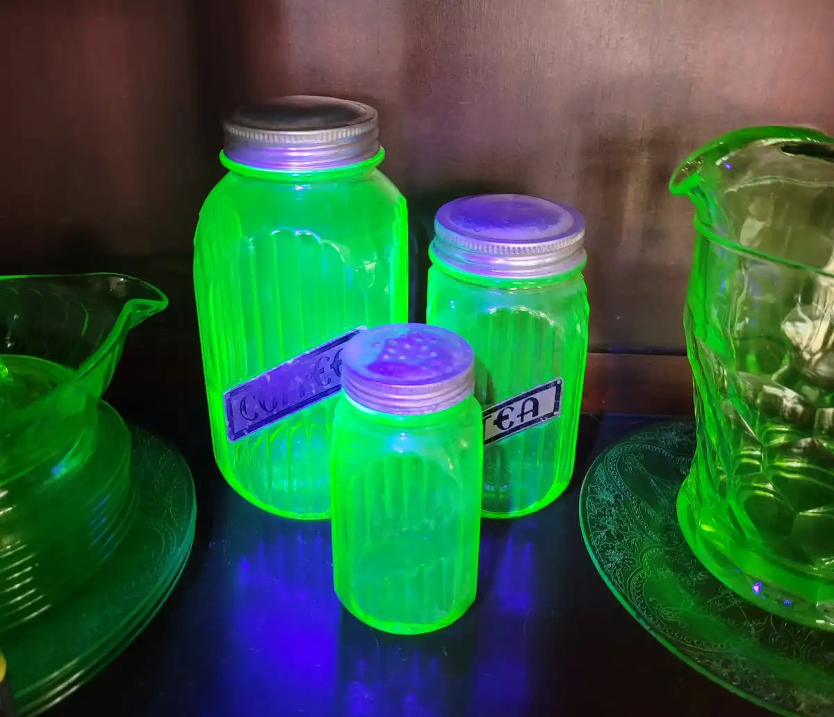 Three glowing green glass jars with metal lids sit on a dark surface under UV light, labeled 'coffee' and 'tea' in reversed…