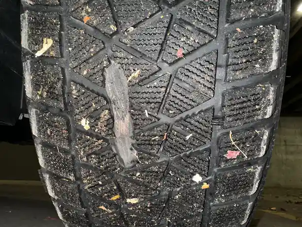A close-up photo shows the tread of a dirty black car tire with scattered leaves and debris, featuring a long, dark stick…