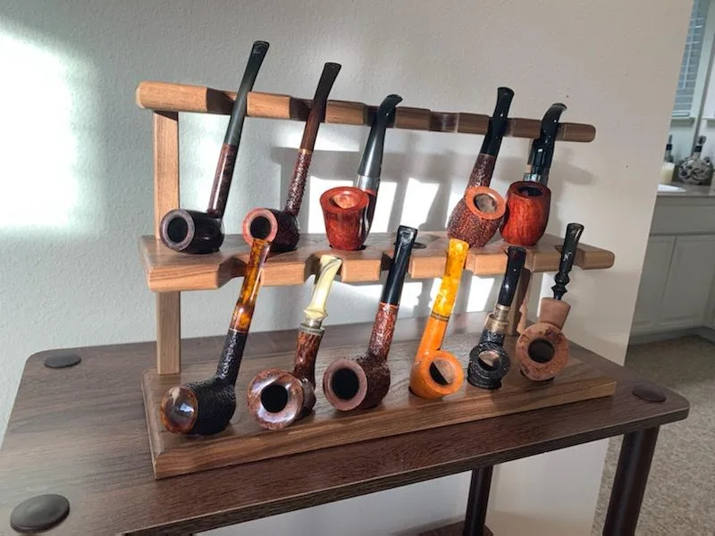 A wooden display rack holds ten smoking pipes of various shapes, colors, and materials, arranged on two tiers on a dark wood…