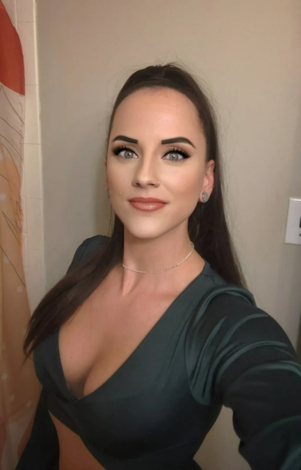 A woman with long dark hair, heavy eyeliner, and a nose ring takes a selfie in a dark green top with a plunging neckline…