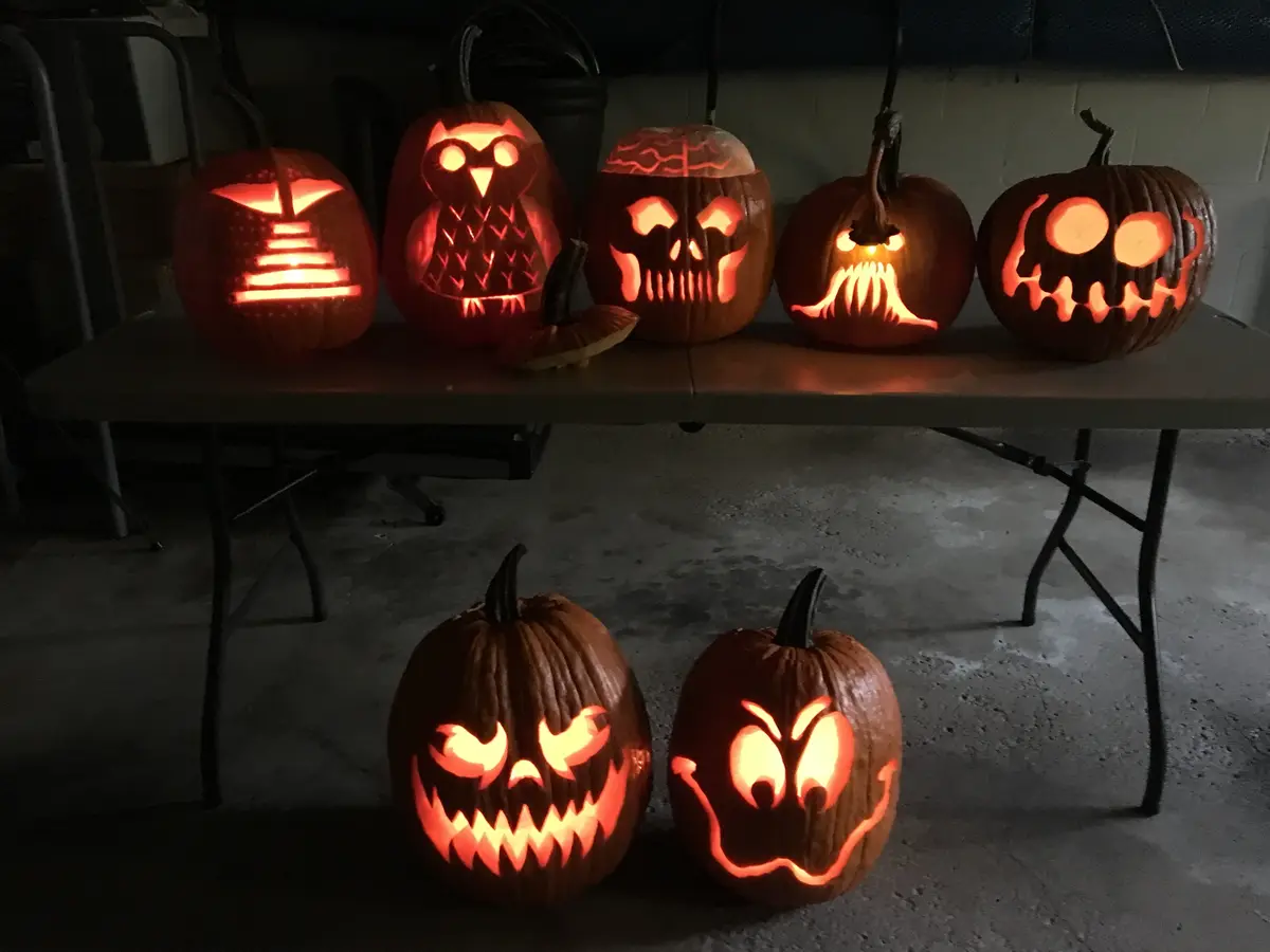 Seven carved pumpkins with glowing orange interiors sit on a folding table and floor in a dimly lit room. The top row…