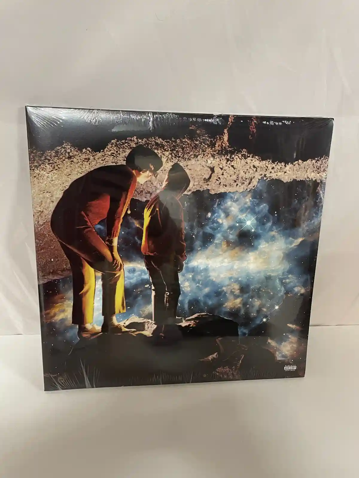 A sealed vinyl record album cover shows two figures standing on a rocky surface, looking down into a cosmic nebula. The…