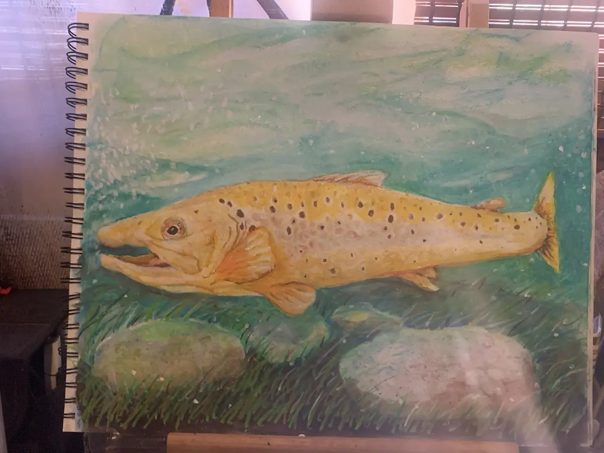 A watercolor painting in a spiral-bound sketchbook depicts a yellowish fish with dark spots swimming in greenish-blue water…