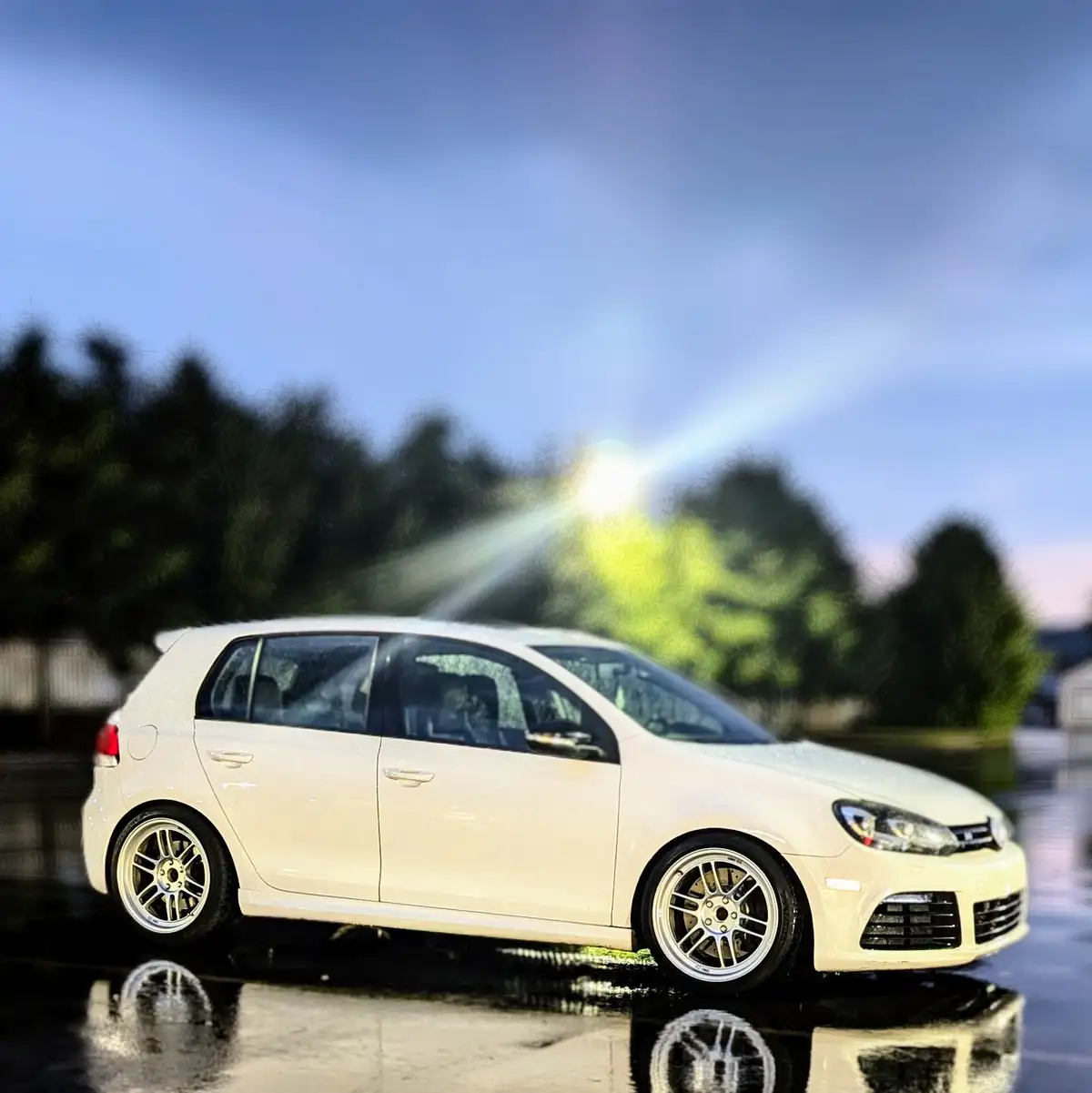 A white Volkswagen Golf is parked on a wet surface, with a bright sunburst effect behind it, creating a dramatic, almost…