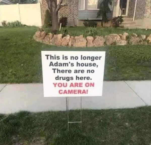 A white yard sign on a metal stand in a grassy front yard reads: 'This is no longer Adam's house, There are no drugs here…