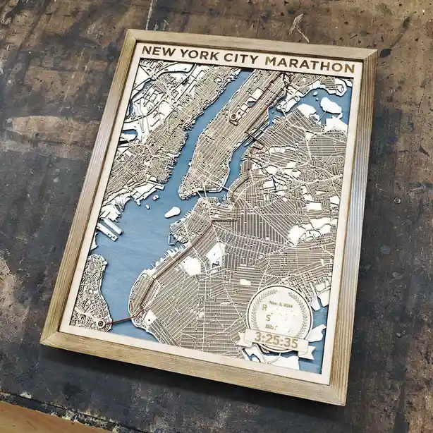 A framed, laser-cut wooden map of New York City shows the marathon route in red, with a circular plaque at the bottom right…