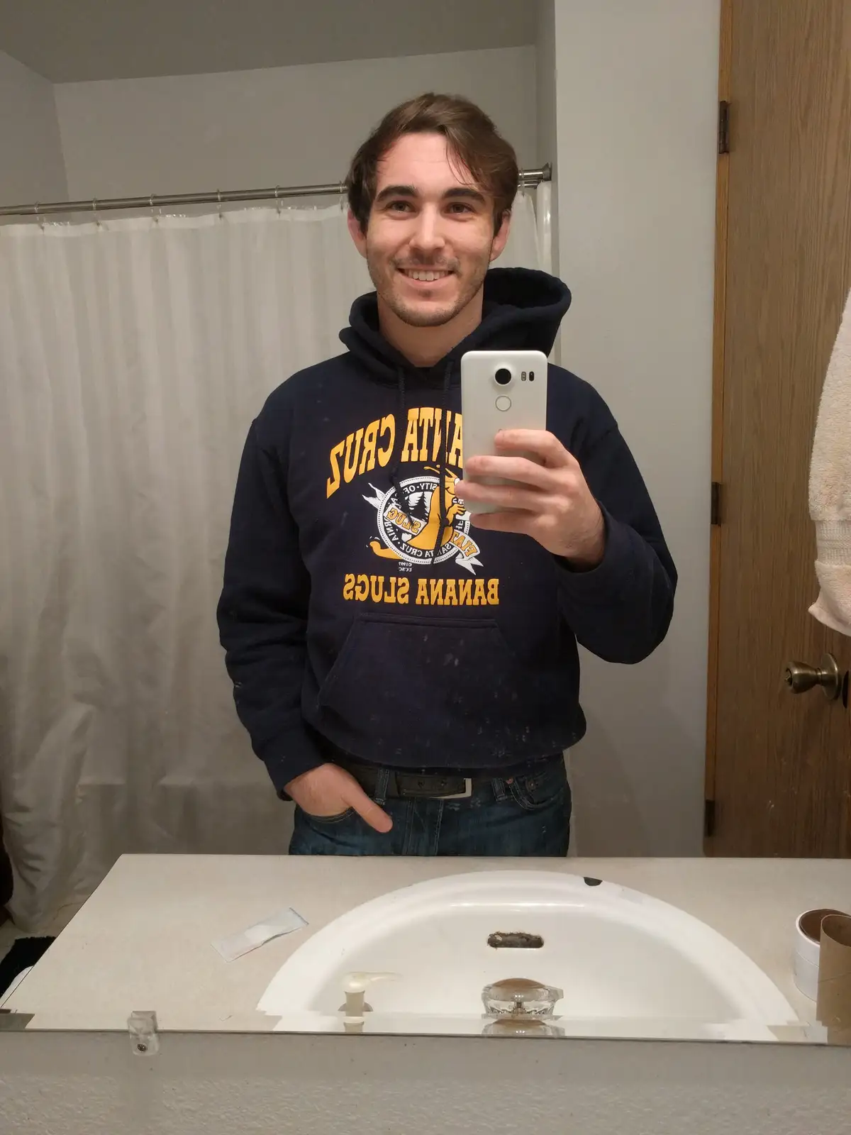 A man in a navy blue 'Santa Cruz Banana Slugs' hoodie takes a mirror selfie in a bathroom, smiling while holding a silver…