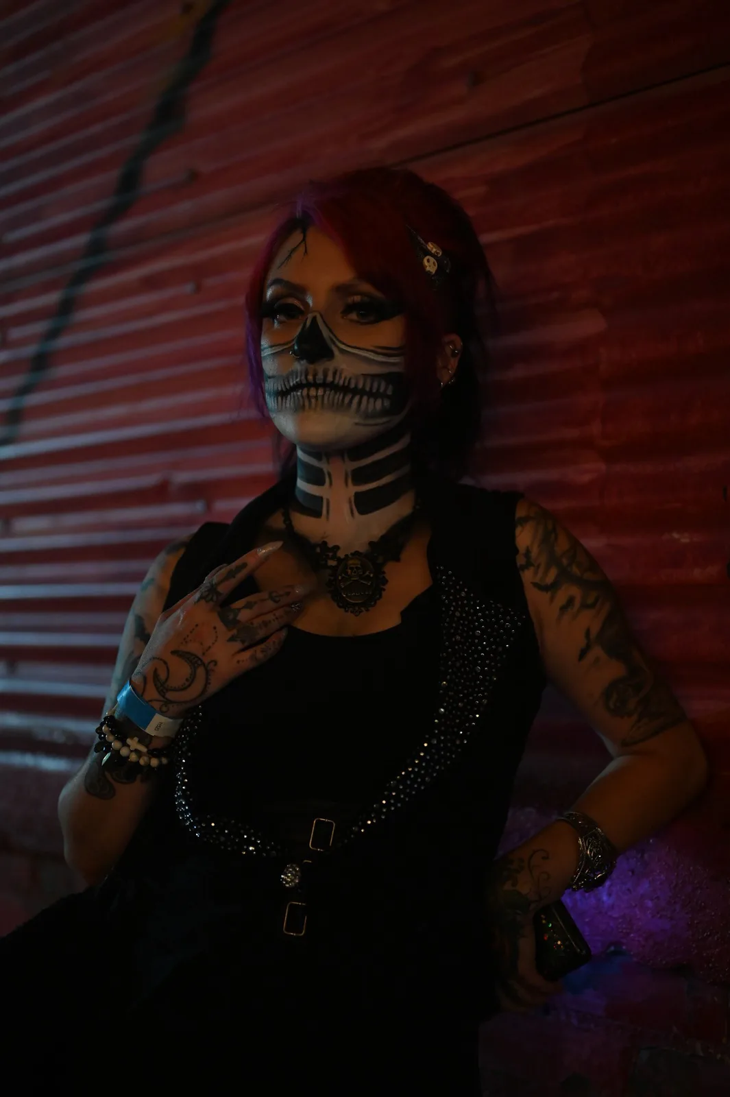 A person with red and purple hair, wearing elaborate skull makeup and black clothing, poses with one hand on their chest…