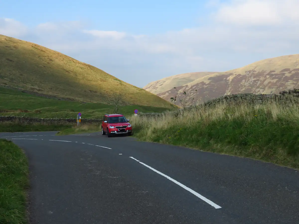 A red car is parked on a narrow rural road flanked by grassy hills and stone walls, with two 'no parking' signs visible…