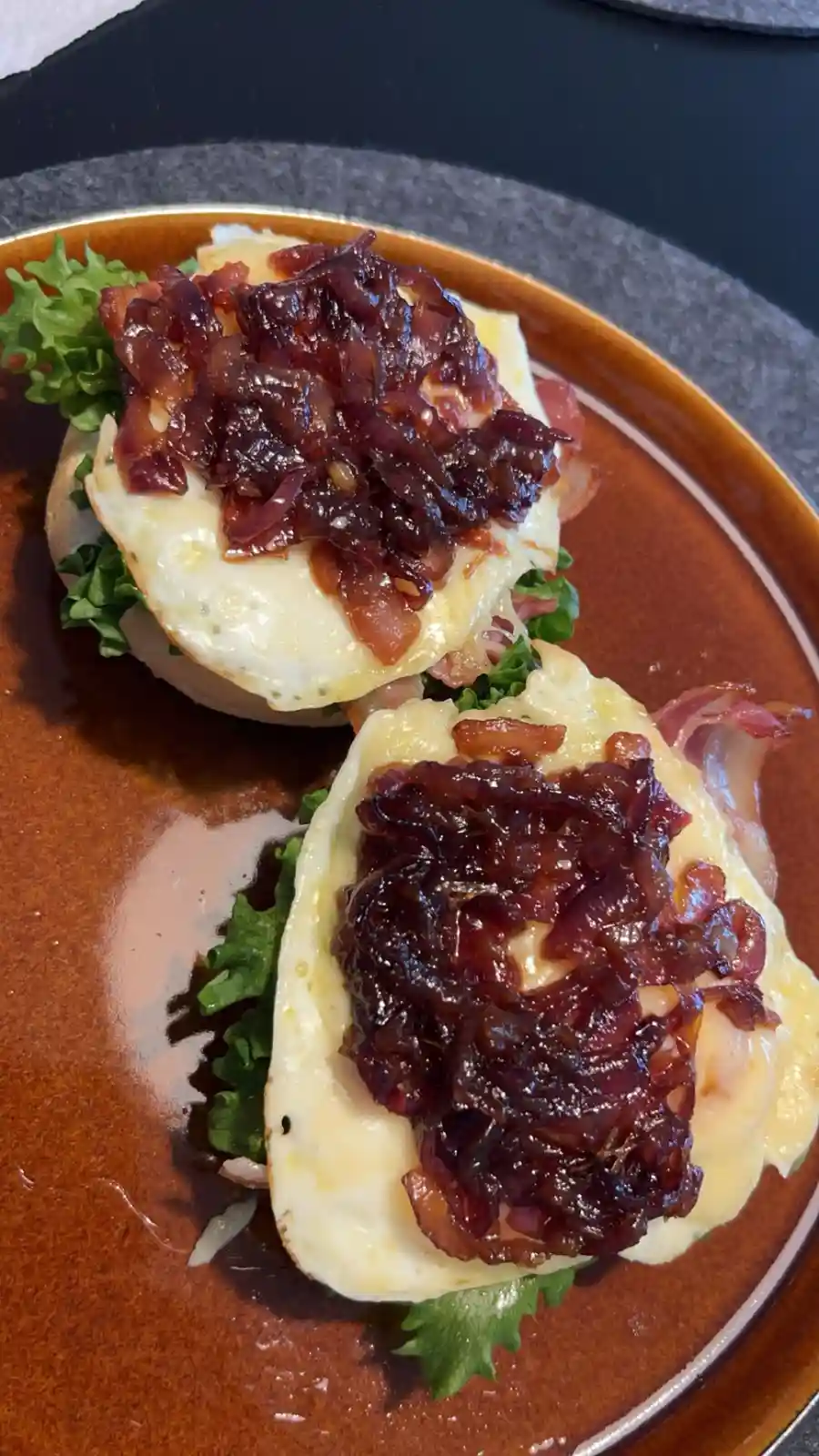 A close-up photo shows two eggs benedict on a brown plate, each with a poached egg, melted cheese, lettuce, and a generous…