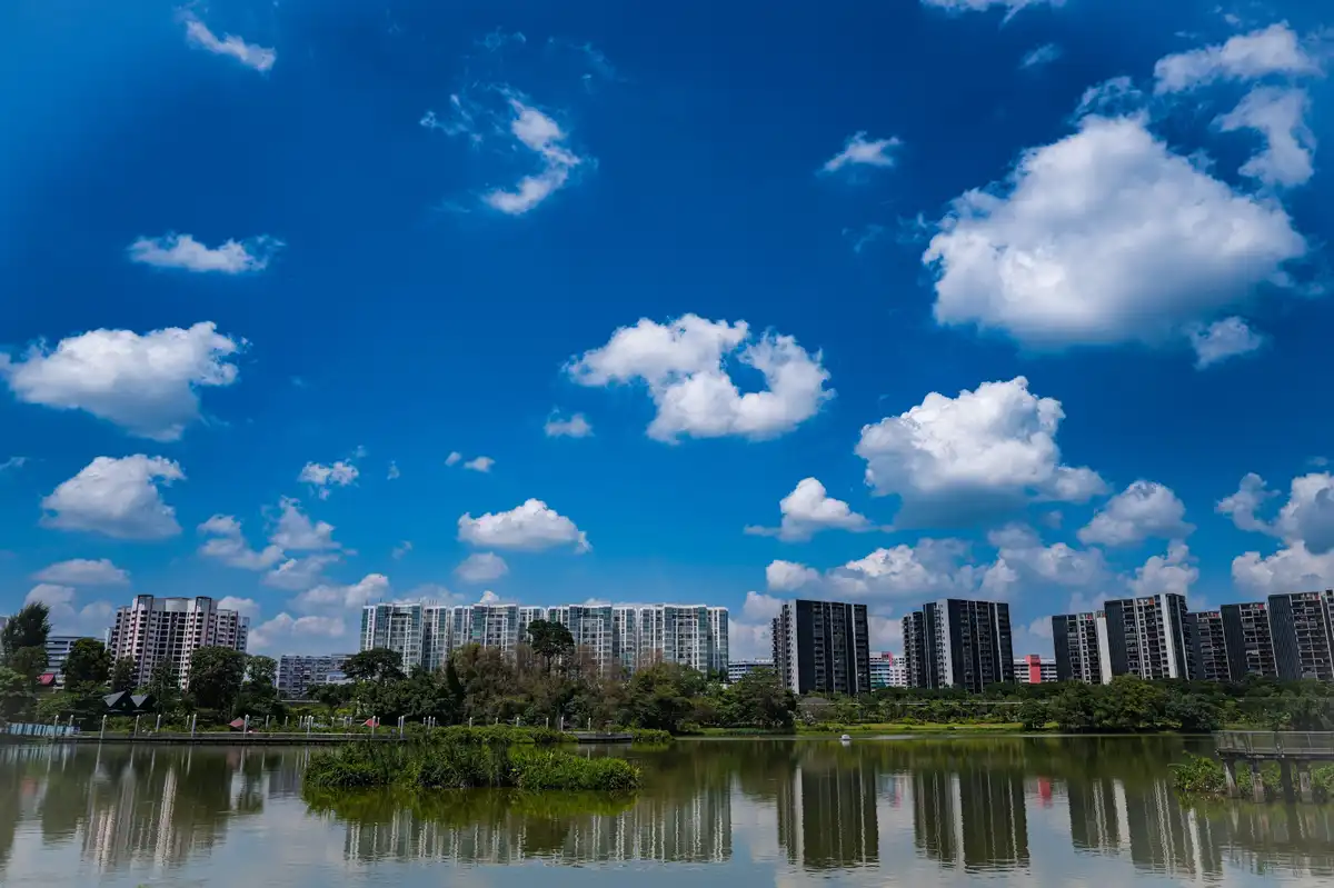 A scenic photo of a calm lake reflecting modern apartment buildings under a bright blue sky with scattered white clouds. One…