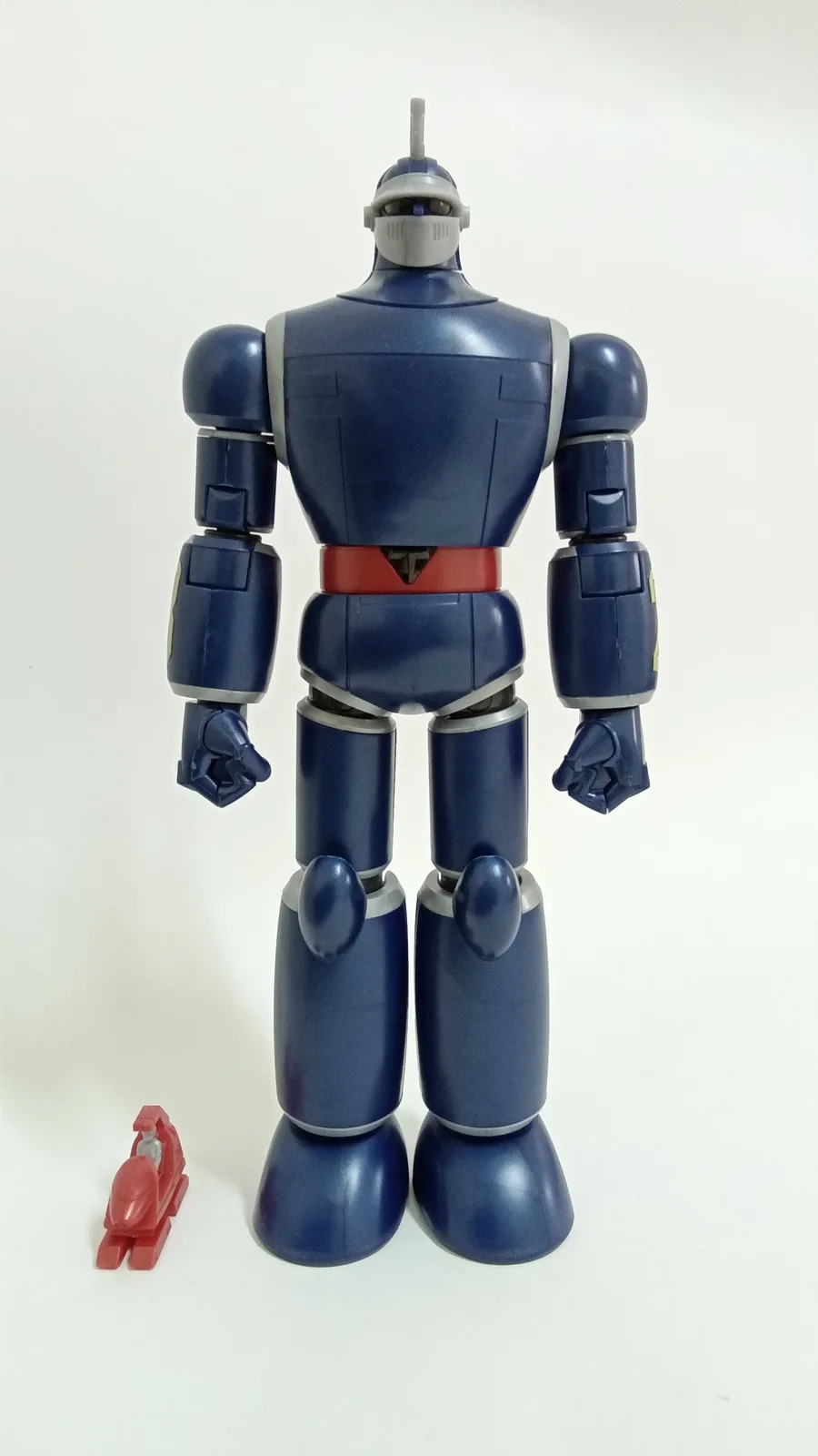 A large blue robot action figure with silver accents and a red belt stands against a white background, with a tiny red toy…
