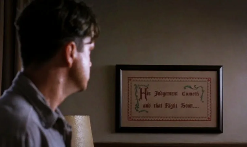 A man in profile looks at a framed piece of embroidery on the wall that reads 'His Judgement Cometh and that Right Soon...'…