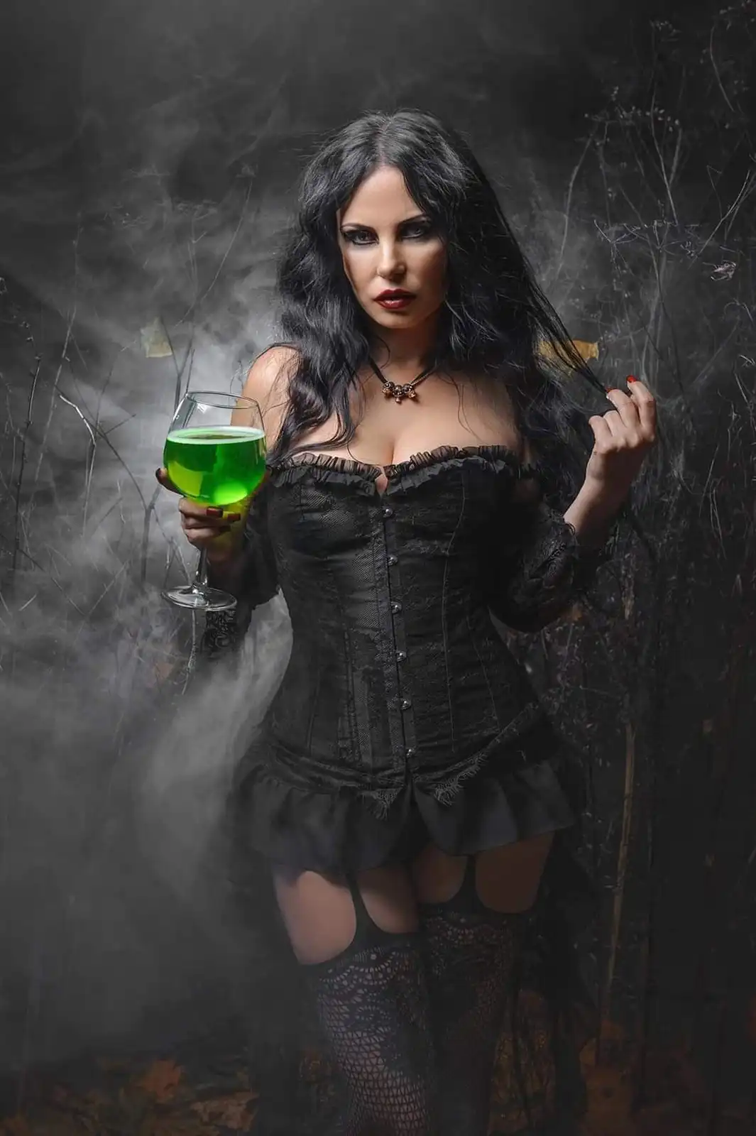 A woman with long black hair, wearing a black corset and fishnet stockings, stands in a smoky, dark setting holding a glass…