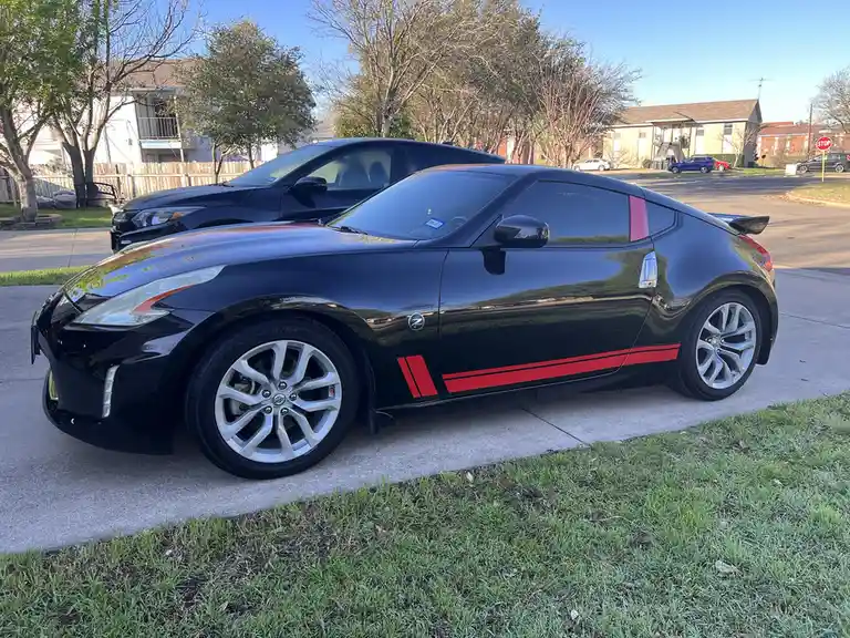 A black Nissan 370Z sports car with red side stripes is parked on a residential driveway, with another dark car behind it…