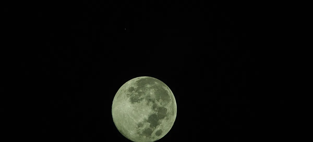 A large, detailed, greenish-tinted full moon dominates a black night sky, with a small, distant star visible above it. The…