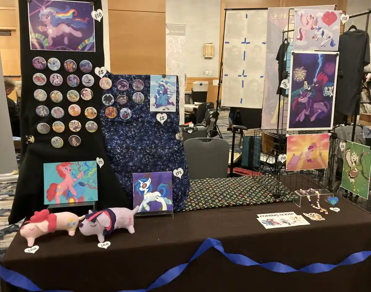 A convention vendor table displays various 'My Little Pony' themed merchandise including paintings, pins, plush toys, and…