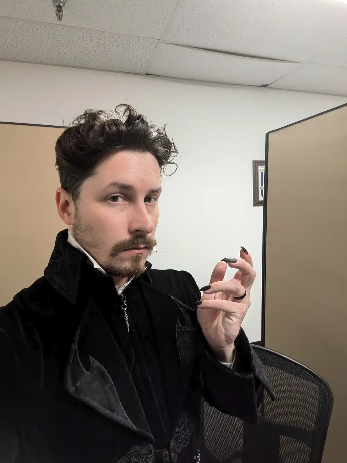 A man with dark, curly hair and a goatee sits in an office chair, wearing a black velvet jacket with bat-wing details and a…