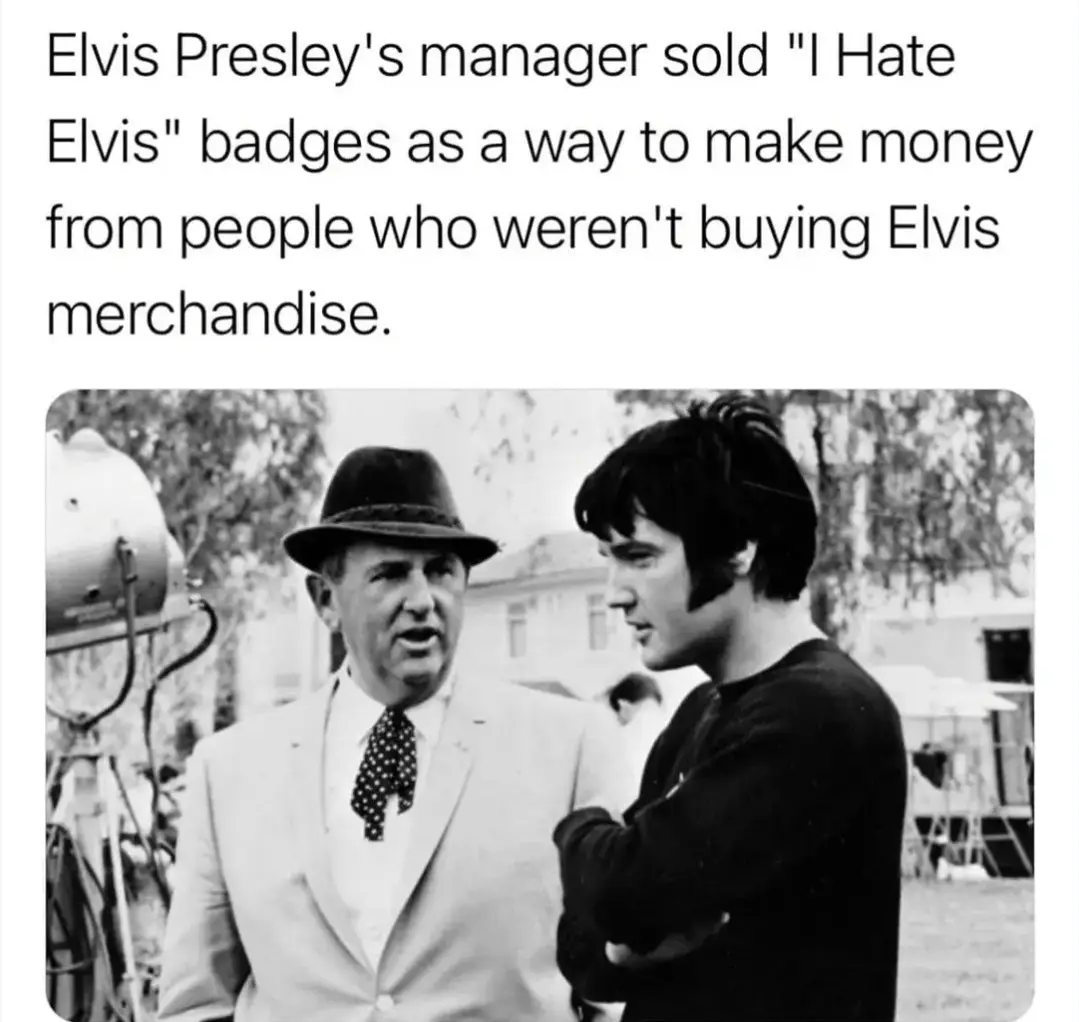 A black-and-white photo shows two men outdoors, one in a light suit and fedora speaking to another man in a dark sweater…