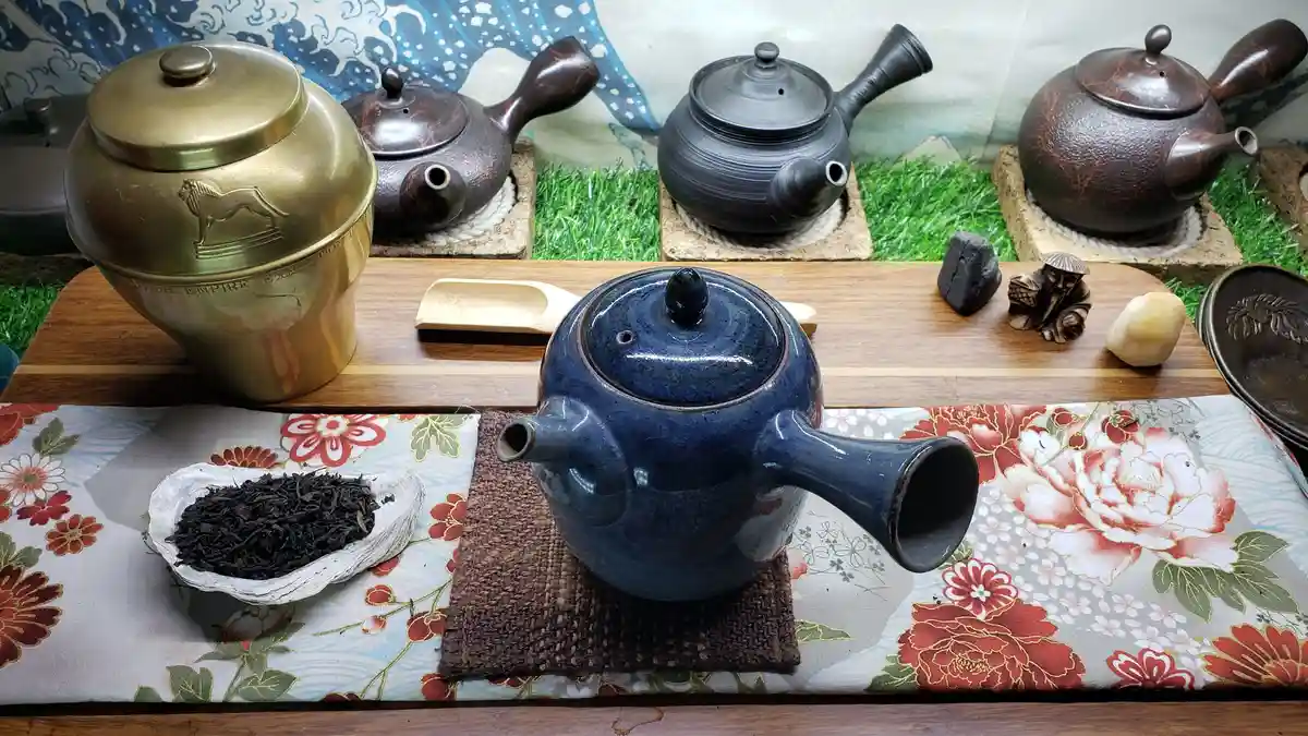 A collection of various teapots and tea-related items arranged on a wooden tray and floral cloth, including a prominent blue…