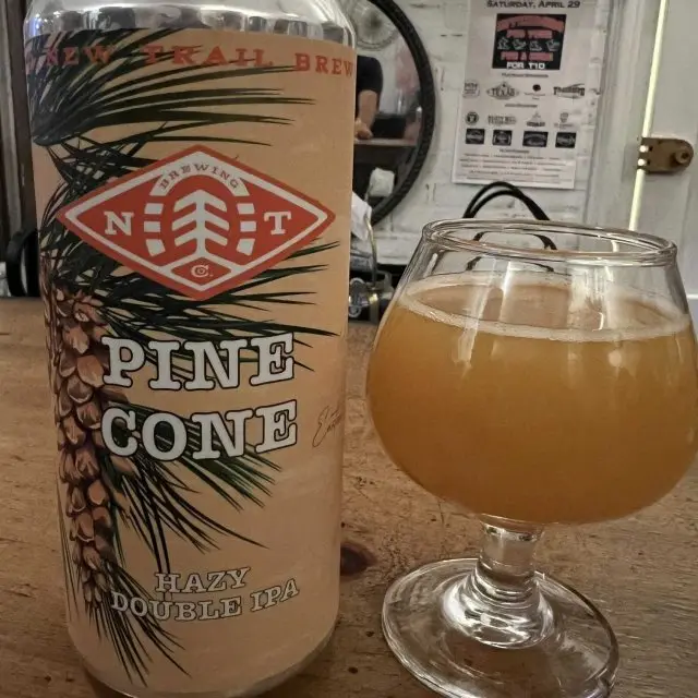 A beer can labeled 'New Trail Brewing Pine Cone Hazy Double IPA' sits next to a glass of the same beer on a wooden table…
