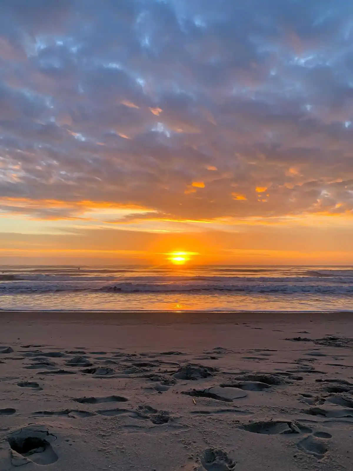 A serene beach sunset with the sun low on the horizon, casting orange and purple hues across the sky and reflecting on the…