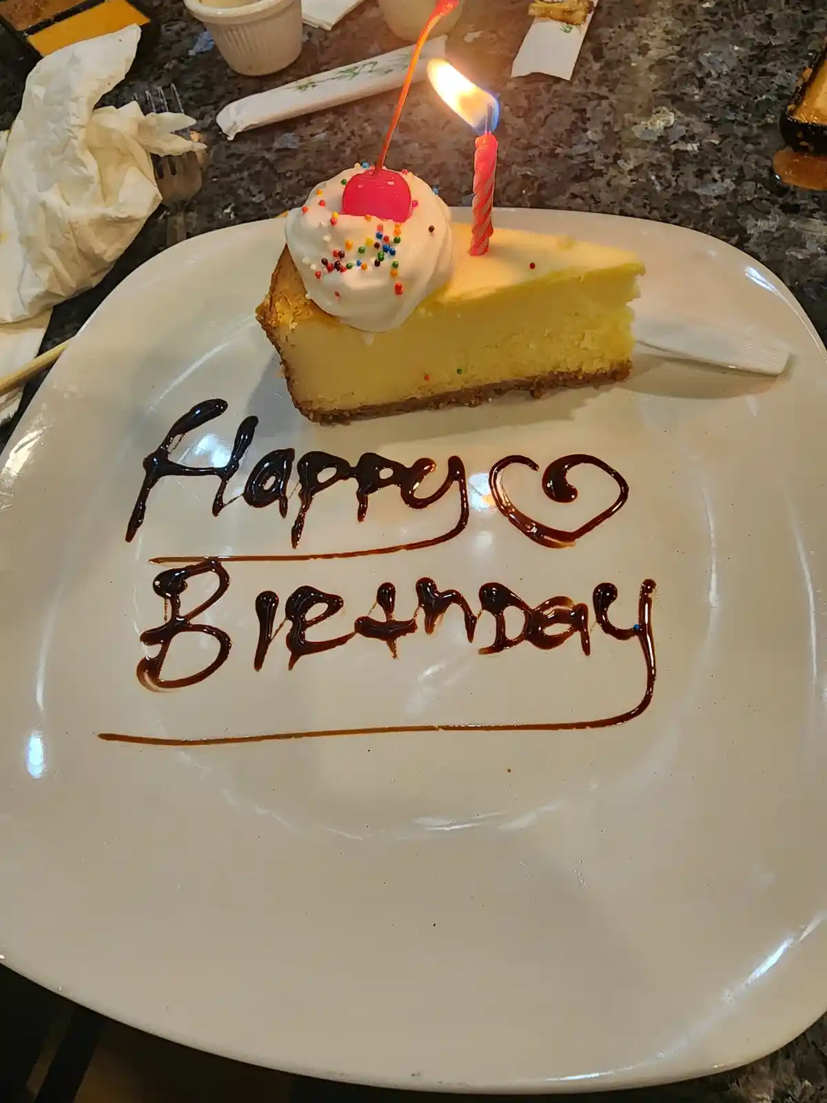 A slice of cheesecake with a lit candle and whipped cream topped with a cherry sits on a white plate. The plate has 'Happy…