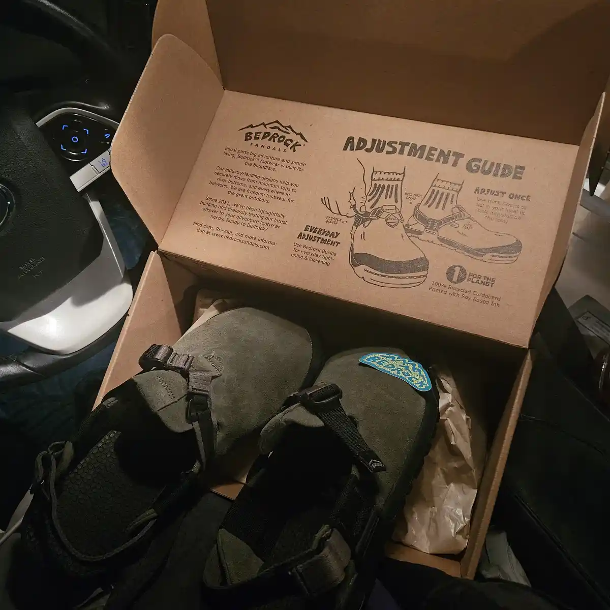 A cardboard box of Bedrock Sandals sits open on a car seat, revealing a pair of olive green sandals with straps. The inside…