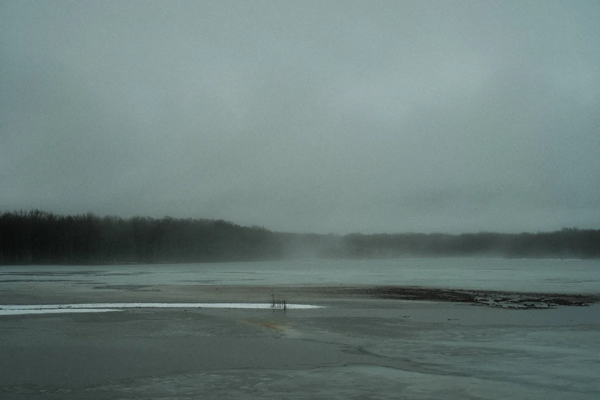 A desolate, foggy landscape shows a frozen or icy body of water with a distant treeline under a gray sky. A small patch of…