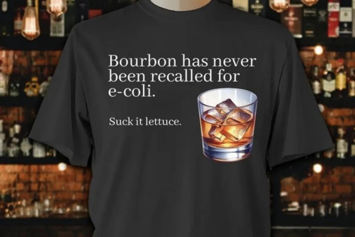 A black t-shirt displays white text stating 'Bourbon has never been recalled for e-coli.' and 'Suck it lettuce.' alongside a…