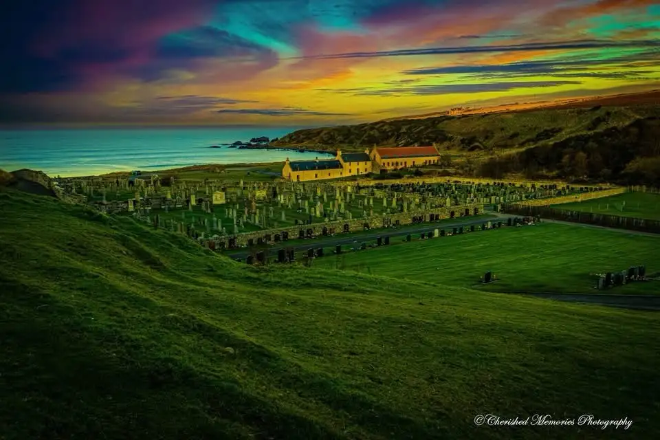 A scenic photograph of a coastal cemetery at sunset, featuring rows of gravestones, a yellow building, and a vibrant sky of…