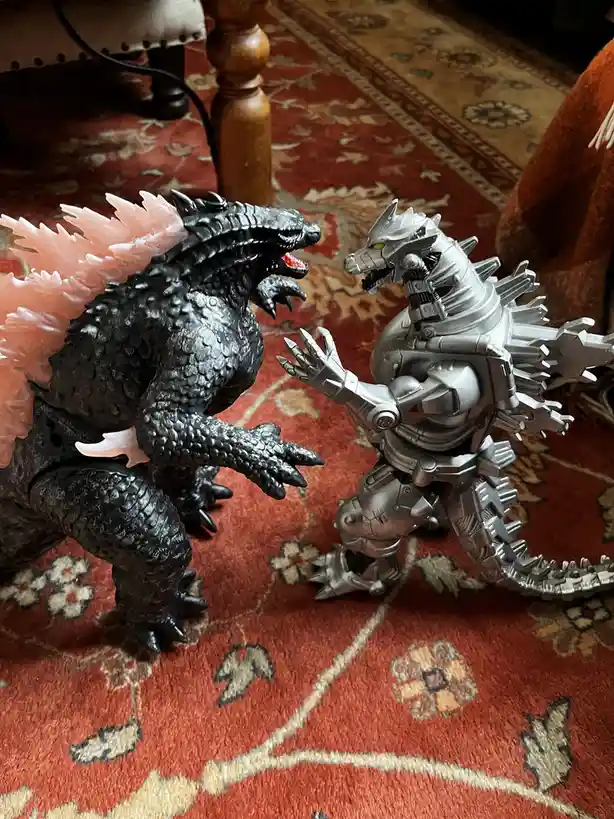 Two Godzilla action figures, one black with pink dorsal fins and one silver robotic version, face each other on a red…
